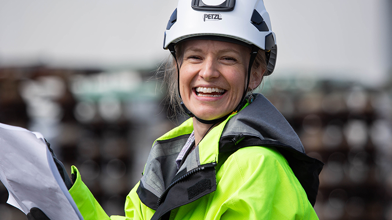 headshot of woman smiling while wearing a hard hat and wearing a bright green coat and holding papers