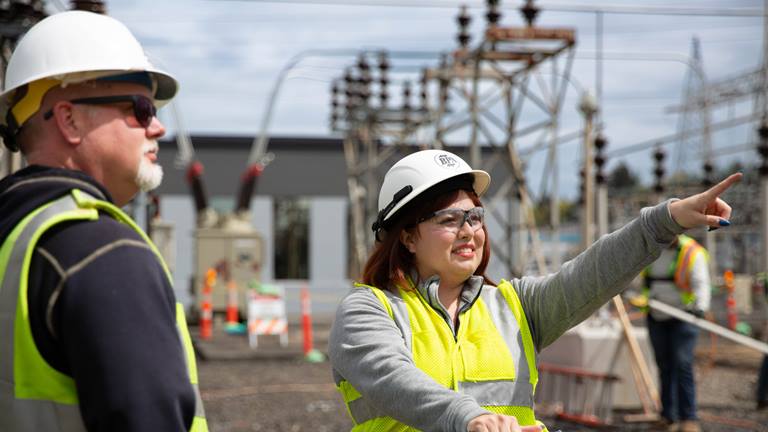 picture of man in hard hat and woman in hard hat, pointing, with transmission structures in the background